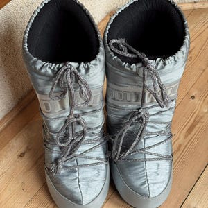 May include: A pair of silver-gray Moon Boots with a shiny, quilted texture. The boots have a lace-up design with gray laces and a black interior. The soles and toe caps are a solid gray color. The boots are designed for cold weather.