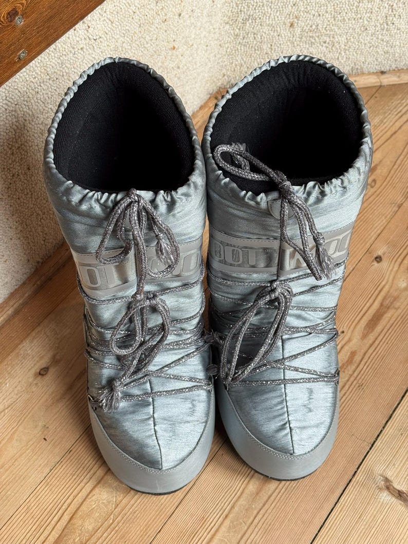 May include: A pair of silver-gray Moon Boots with a shiny, quilted texture. The boots have a lace-up design with gray laces and a black interior. The soles and toe caps are a solid gray color. The boots are designed for cold weather.