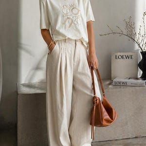 May include: Cream-colored t-shirt with the "LOEWE" logo, paired with wide-leg, light-colored pants. A brown leather handbag and white sneakers complete the outfit. A book with the "LOEWE" logo is visible in the background.