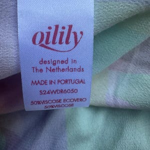 Pu&ograve; includere: Primo piano di un'etichetta di tessuto con il marchio "oilily" in corsivo bordeaux. L'etichetta recita "designed in The Netherlands" e "MADE IN PORTUGAL". Il tessuto presenta un motivo a quadri pastello nei toni del viola, verde e blu.
