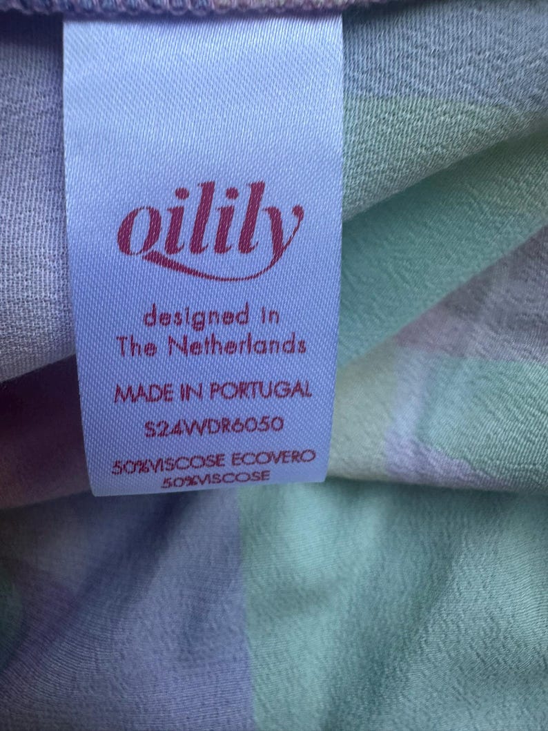 Pu&ograve; includere: Primo piano di un'etichetta di tessuto con il marchio "oilily" in corsivo bordeaux. L'etichetta recita "designed in The Netherlands" e "MADE IN PORTUGAL". Il tessuto presenta un motivo a quadri pastello nei toni del viola, verde e blu.