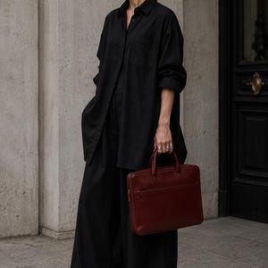 May include: A black button-down shirt with long sleeves and a chest pocket, paired with loose-fitting black trousers. The ensemble is accessorized with a dark burgundy leather briefcase featuring dual top handles and a front zippered compartment.