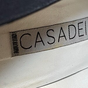 May include: Close-up of a shoe interior with a label. The label reads "CASADEI" in black capital letters on a gray fabric. The background is white leather, with a hint of blue at the top.