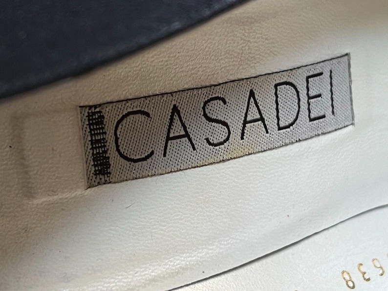 May include: Close-up of a shoe interior with a label. The label reads "CASADEI" in black capital letters on a gray fabric. The background is white leather, with a hint of blue at the top.