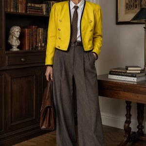 Könnte beinhalten: Eine leuchtend gelbe, kurz geschnittene Jacke mit schwarzer Umrandung und goldenen Knöpfen, getragen über einem weißen Hemd mit brauner Krawatte. Dazu gehören eine dunkelbraune, hoch taillierte Hose mit weitem Bein, ein Ledergürtel und eine Ledertasche.