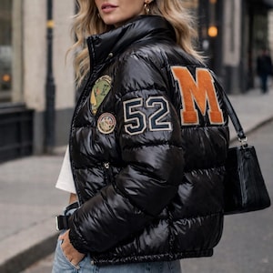 Könnte beinhalten: Schwarze Steppjacke mit der Zahl "52" und dem Buchstaben "M" in Orange auf dem Rücken. Die Jacke hat einen hohen Kragen und mehrere Aufnäher. Die Person trägt eine Sonnenbrille und hält eine schwarze Handtasche.