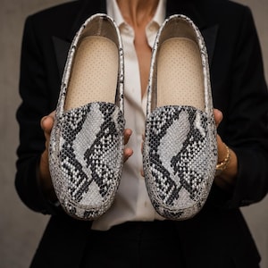 Könnte beinhalten: Ein Paar Slipper mit einem grau-schwarz-weißen Schlangenledermuster. Die Schuhe haben eine abgerundete Zehenpartie, sichtbare Nähte entlang des Vorderblatts und ein helles Innenfutter mit einer perforierten Textur auf der Innensohle.