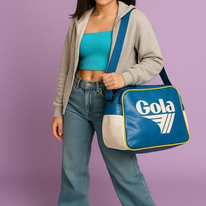 May include: A person wearing a teal crop top, gray zip-up hoodie, and blue jeans, carrying a blue and white Gola shoulder bag. The bag has the Gola logo in white. The background is a solid purple.