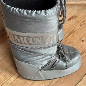 May include: Silver-colored Moon Boot winter boots. The boots have a shiny, satin-like finish with the brand name printed on a band around the mid-section. They feature gray laces and a black interior lining.
