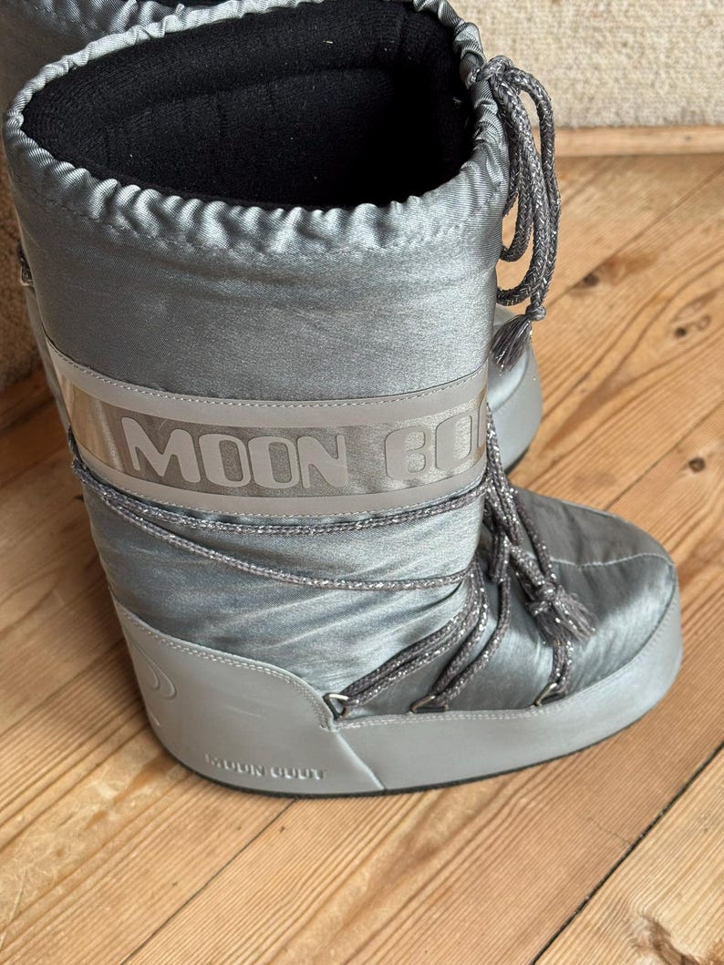 May include: Silver-colored Moon Boot winter boots. The boots have a shiny, satin-like finish with the brand name printed on a band around the mid-section. They feature gray laces and a black interior lining.