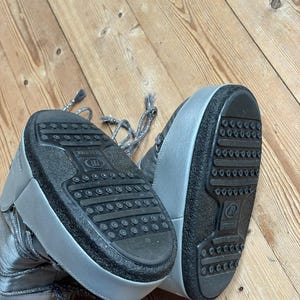 May include: Silver-colored winter boots with black soles. The boots have a high-top design with laces and the word "MOON" visible on the side. The soles have a textured pattern for grip.