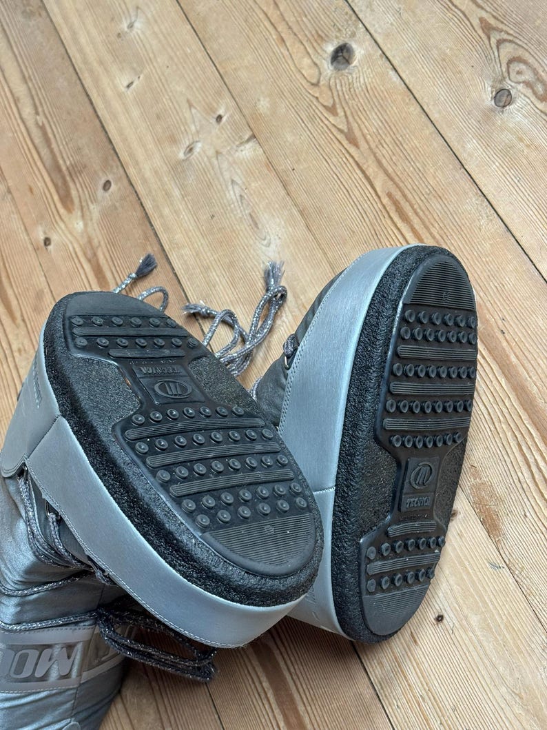 May include: Silver-colored winter boots with black soles. The boots have a high-top design with laces and the word "MOON" visible on the side. The soles have a textured pattern for grip.