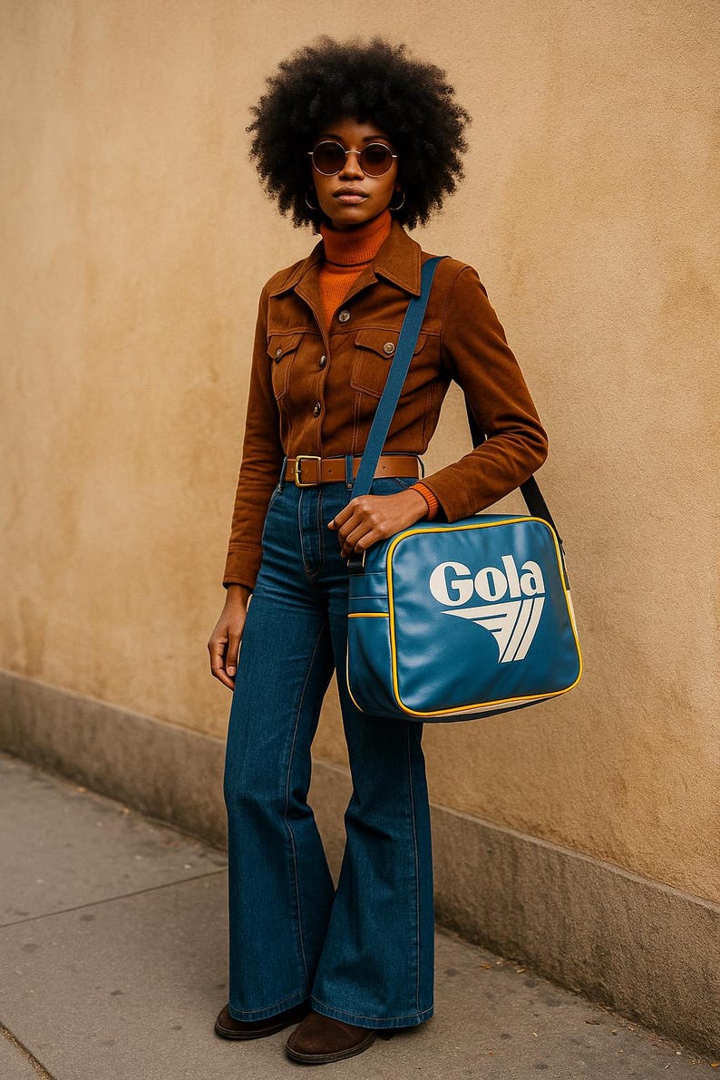 May include: A person wearing a brown suede jacket, orange turtleneck, and flared blue jeans. A blue shoulder bag with the white text "Gola" is visible. The outfit is accessorized with a brown belt and sunglasses. The background is a light brown wall.
