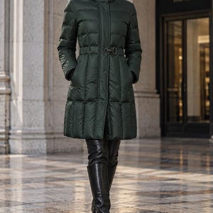 May include: A dark green, knee-length, quilted puffer coat featuring a high collar, a front zipper closure, and a metal buckle detail at the waist. The coat has long sleeves and a smooth, water-resistant finish, providing a structured silhouette.