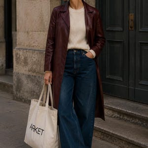K&ouml;nnte beinhalten: Ein langer bordeauxroter Ledermantel &uuml;ber einem cremefarbenen Strickpullover und einer dunkelblauen, weit geschnittenen Jeans. Das Outfit umfasst wei&szlig;e Sneaker und eine beige Stofftasche mit dem Wort ARKET in schwarzem Druck auf der Vorderseite.