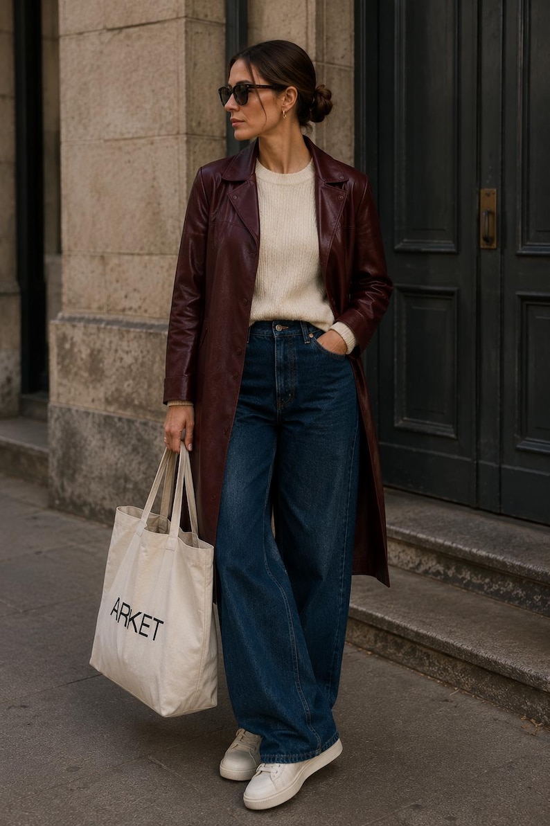 K&ouml;nnte beinhalten: Ein langer bordeauxroter Ledermantel &uuml;ber einem cremefarbenen Strickpullover und einer dunkelblauen, weit geschnittenen Jeans. Das Outfit umfasst wei&szlig;e Sneaker und eine beige Stofftasche mit dem Wort ARKET in schwarzem Druck auf der Vorderseite.
