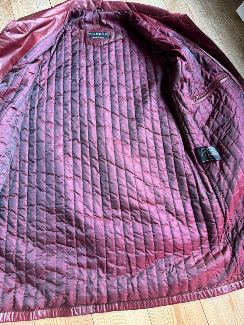K&ouml;nnte beinhalten: Eine bordeauxrote Lederjacke mit gestepptem Innenfutter. Das Innenetikett tr&auml;gt den Text MORENA ART OF FASHION. Die Jacke verf&uuml;gt &uuml;ber ein vertikales Steppmuster auf der Innenseite, eine kleine Innentasche und ein schwarzes Pflegeetikett.
