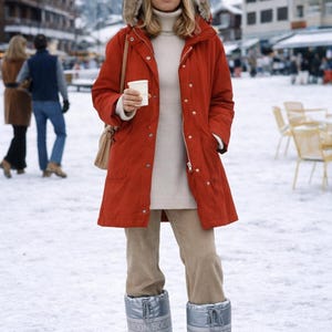 May include: A person wearing a red winter coat with a fur-lined hood, sunglasses, and silver Moon Boots. The person is holding a white cup and standing in a snowy outdoor setting. The outfit includes a cream-colored turtleneck and beige pants.