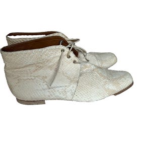 Könnte beinhalten: Ein Paar cremefarbene Stiefeletten mit Schlangenmuster. Die Stiefel haben ein Schnürdesign und einen niedrigen Absatz. Das Innenfutter ist braun. Die Schuhe sind vor weißem Hintergrund.