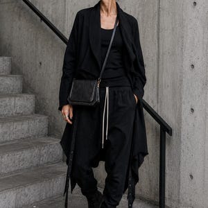 Può includere: Un completo nero composto da una lunga giacca drappeggiata aperta sul davanti con bordi sfrangiati, una canotta nera e pantaloni da jogging neri con una lunga coulisse chiara. Una borsa a tracolla nera, stivali con plateau e occhiali da sole.