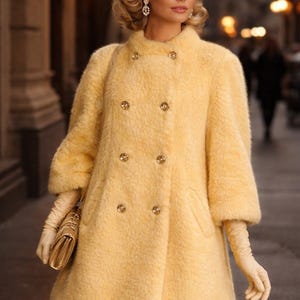 Könnte beinhalten: Ein hellgelber, doppelreihiger Mantel mit großen goldenen Knöpfen. Der Mantel hat ein strukturiertes, flauschiges Aussehen und dreiviertel Ärmel. Das Outfit wird durch passende Handschuhe und eine goldene Handtasche ergänzt. Der Mantel ist knielang.