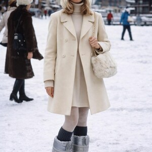 May include: A woman in a cream-colored coat, dress, and sunglasses stands in the snow. She wears silver snow boots and carries a fluffy purse. The background shows a snowy landscape with buildings and other people.