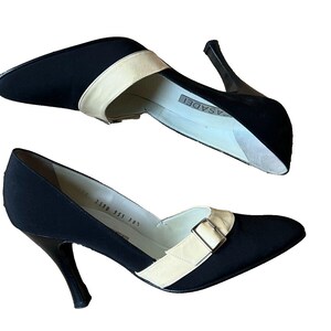May include: Pair of black pumps with a pointed toe and stiletto heel. The shoes feature a cream-colored strap across the top with a silver buckle. The heels are approximately 3 inches (7.6 cm) high.