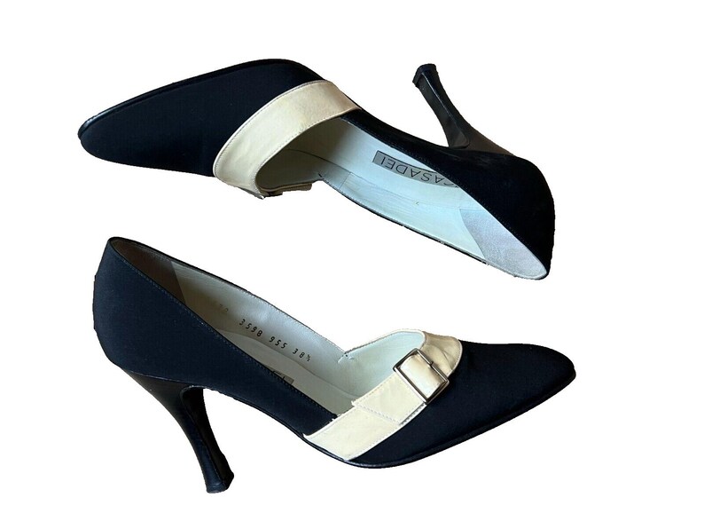 May include: Pair of black pumps with a pointed toe and stiletto heel. The shoes feature a cream-colored strap across the top with a silver buckle. The heels are approximately 3 inches (7.6 cm) high.