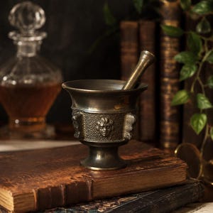 Può includere: Un mortaio e un pestello in bronzo con dettagliate sculture di volti sono appoggiati su una pila di libri antichi. Il mortaio è parzialmente riempito con una sostanza scura e un pestello è all'interno. Una caraffa di vetro e vecchi libri sono sullo sfondo.