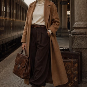 May include: A long camel-colored wool coat worn over a cream turtleneck sweater and dark brown trousers with a brown leather belt. The outfit includes a brown leather briefcase and lace-up boots with a patterned print, featuring a low heel.