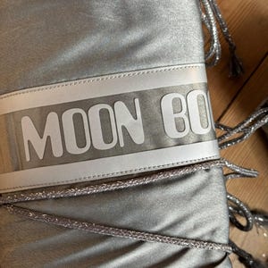 May include: Silver Moon Boot with the brand name in white block letters across the top. The boots have silver drawstrings and a shiny, metallic finish. The boots are designed for cold weather.