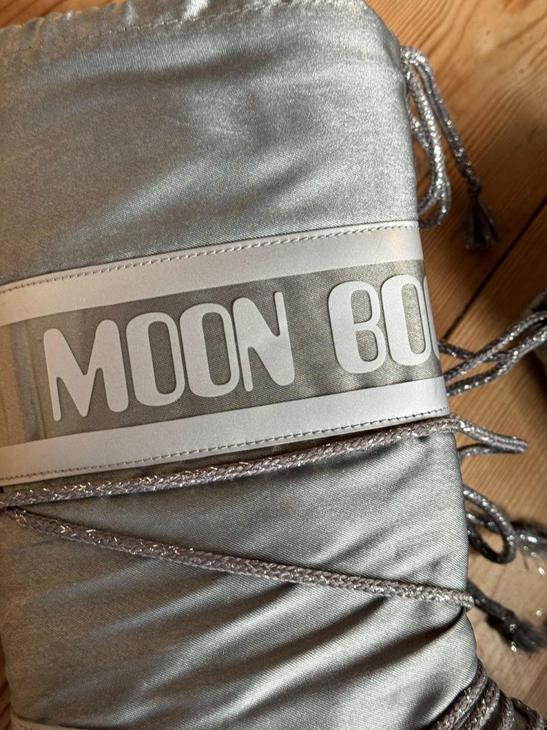 May include: Silver Moon Boot with the brand name in white block letters across the top. The boots have silver drawstrings and a shiny, metallic finish. The boots are designed for cold weather.