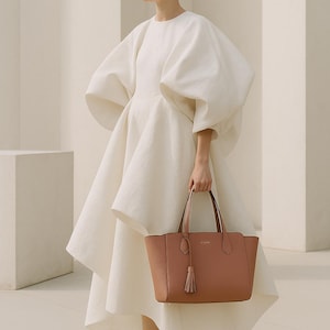 May include: A woman in a white dress with large sleeves and a flared skirt holds a brown leather tote bag with a tassel. She wears white heels. The background is a minimalist, light-colored space.