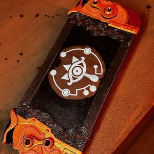 May include: A black Nintendo Switch case with a gold and red design. The case features a stylized eye with a white outline and a circular pattern around it.