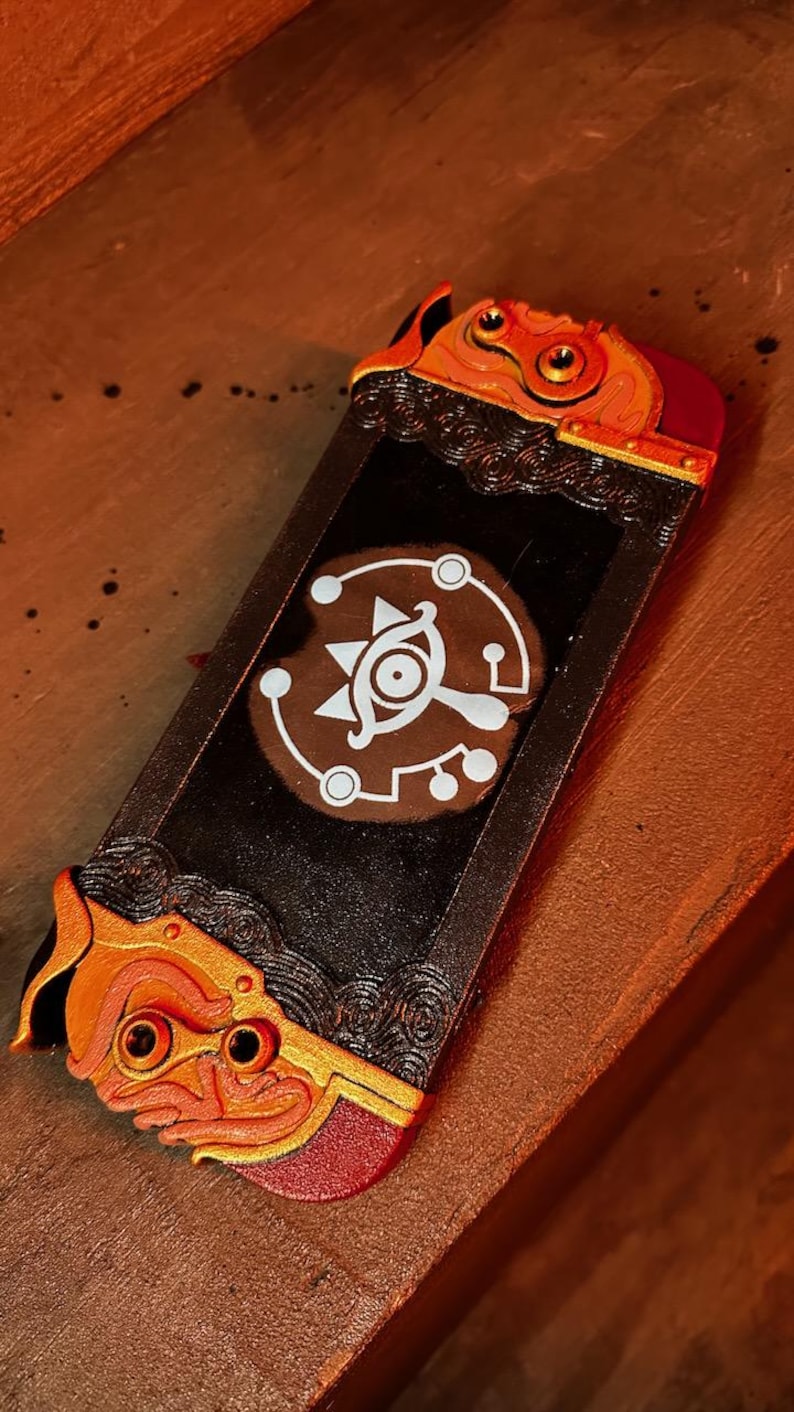 May include: A black Nintendo Switch case with a gold and red design. The case features a stylized eye with a white outline and a circular pattern around it.