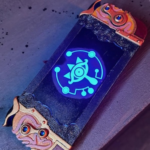 May include: A custom Nintendo Switch console with a black and gold design featuring a glowing blue Triforce symbol. The console has a detailed, intricate design with gold accents.