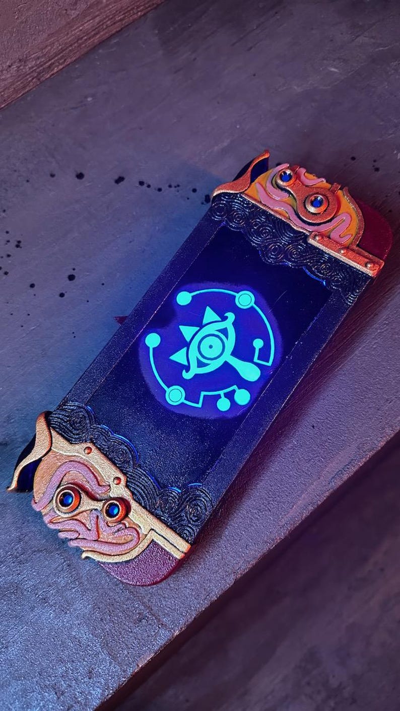 May include: A custom Nintendo Switch console with a black and gold design featuring a glowing blue Triforce symbol. The console has a detailed, intricate design with gold accents.