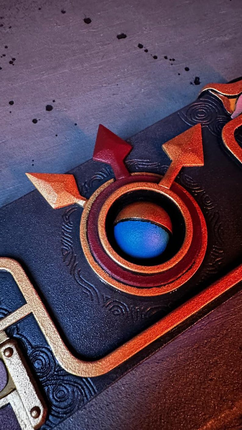 May include: A close-up of a dark blue leather-bound journal with a gold and red decorative clasp. The clasp features a circular design with a blue eye in the center.
