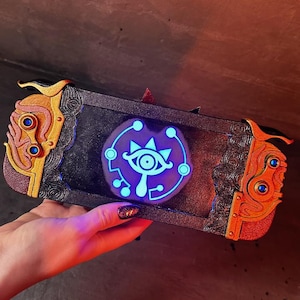 May include: A black Nintendo Switch console with a custom design featuring a gold and orange trim and a glowing blue Sheikah Eye symbol.