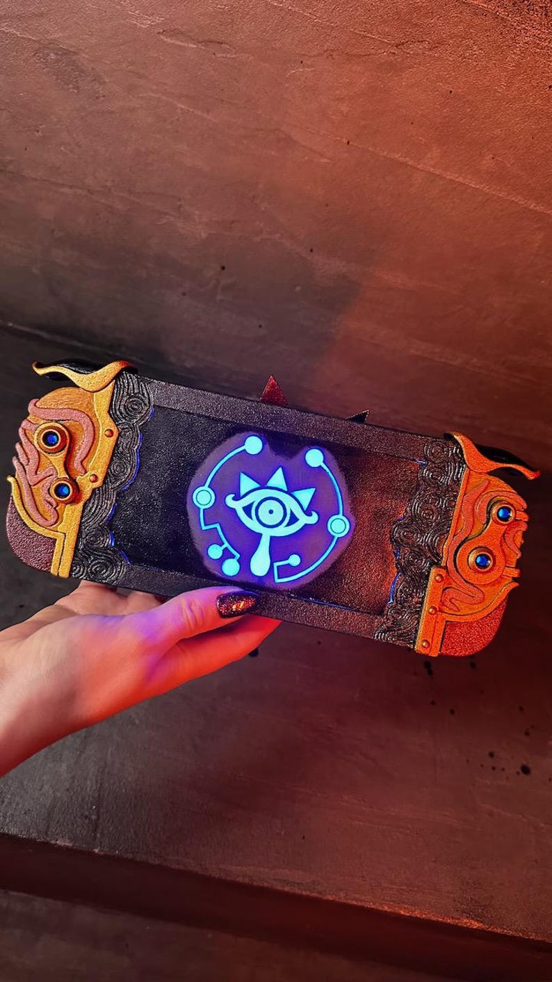 May include: A black Nintendo Switch console with a custom design featuring a gold and orange trim and a glowing blue Sheikah Eye symbol.