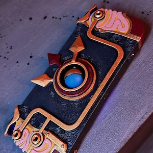 May include: A black and gold phone case with a detailed design featuring a blue and red circle in the center. The case has a steampunk aesthetic with intricate details and a textured surface.