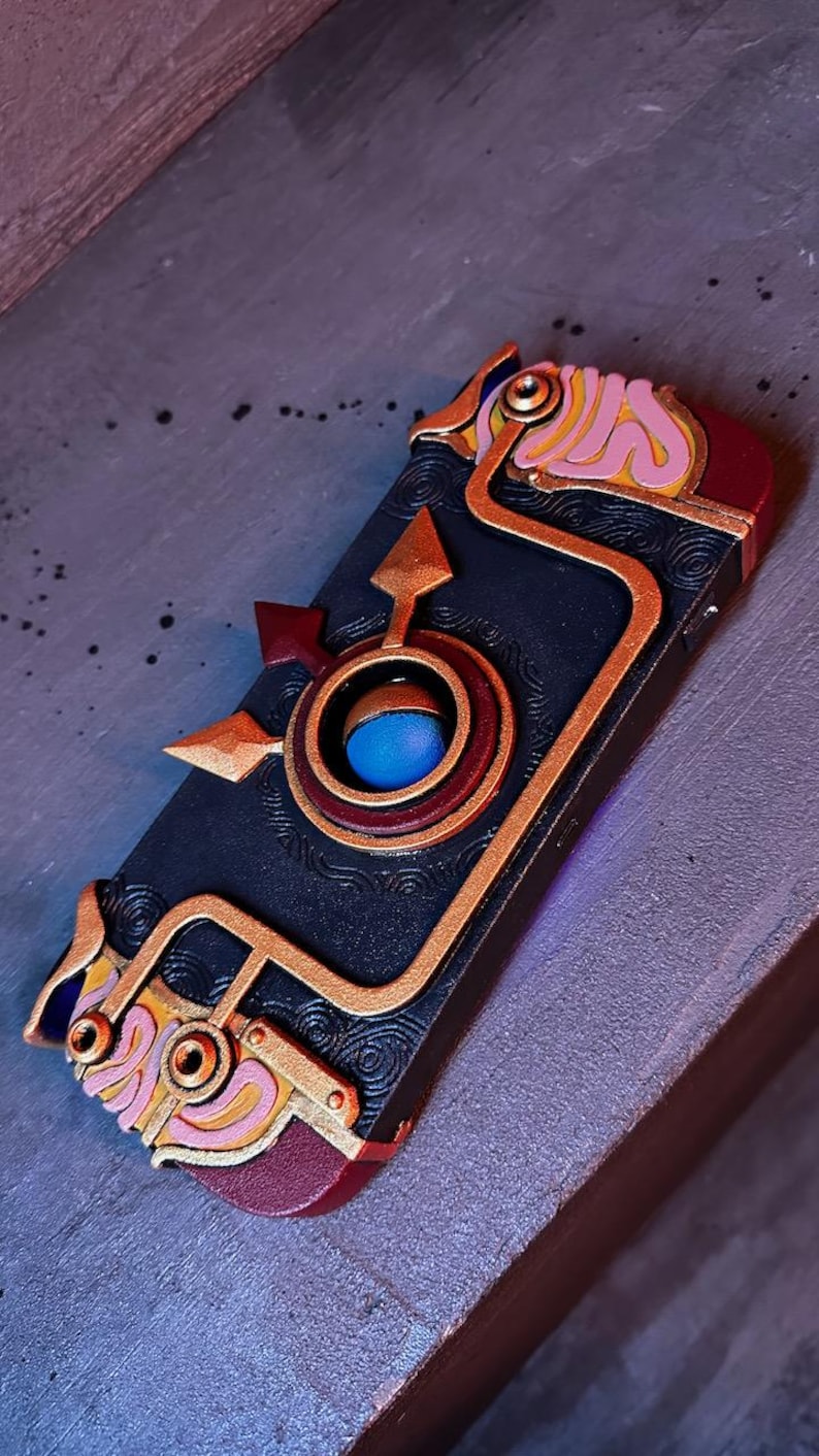 May include: A black and gold phone case with a detailed design featuring a blue and red circle in the center. The case has a steampunk aesthetic with intricate details and a textured surface.