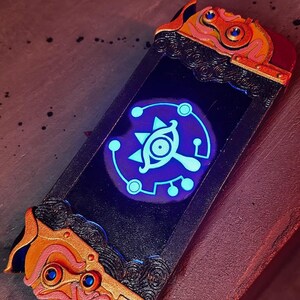 May include: A custom Nintendo Switch controller with a black and gold design. The controller features a glowing blue Triforce symbol with a stylized eye in the center.