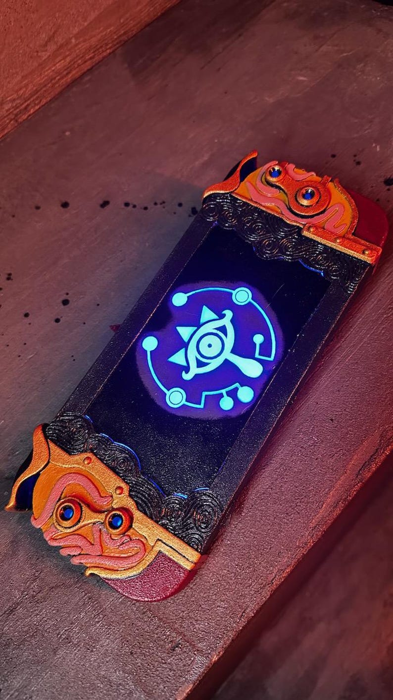 May include: A custom Nintendo Switch controller with a black and gold design. The controller features a glowing blue Triforce symbol with a stylized eye in the center.