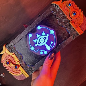 May include: A black Nintendo Switch case with a gold and red trim. The case features a glowing blue Triforce symbol with an eye in the center.