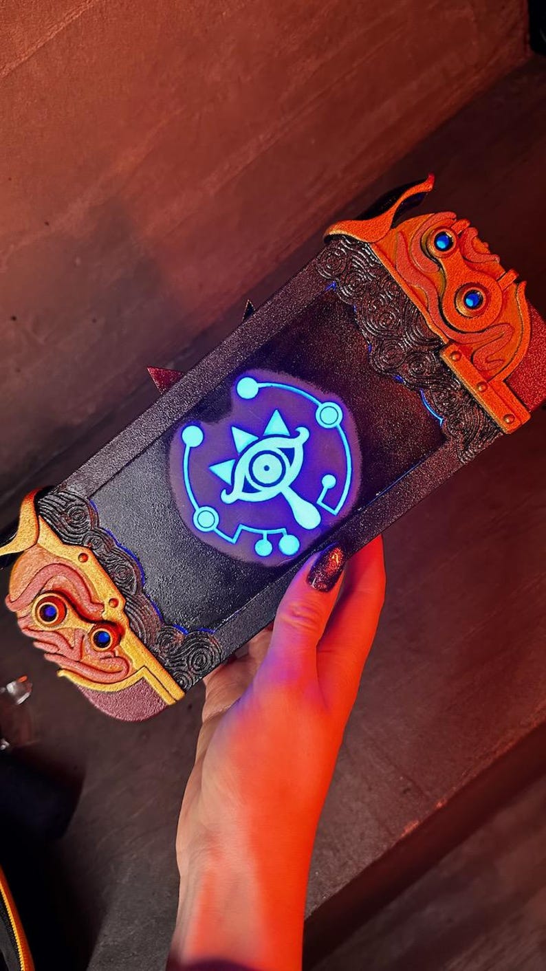 May include: A black Nintendo Switch case with a gold and red trim. The case features a glowing blue Triforce symbol with an eye in the center.