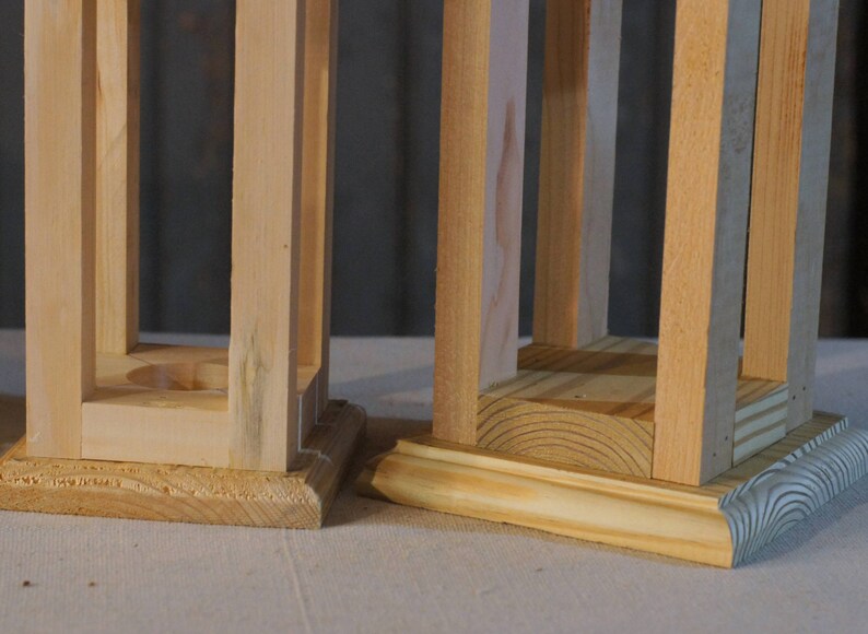 May include: Two unfinished wooden lanterns with square bases. The lantern on the left has a simple base, while the lantern on the right has a more decorative base.
