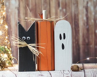 Può includere: Tre decorazioni di Halloween in legno: un gatto nero con occhietti, una zucca arancione e un fantasma bianco con occhi e bocca neri. Le decorazioni sono su una superficie di legno bianca.