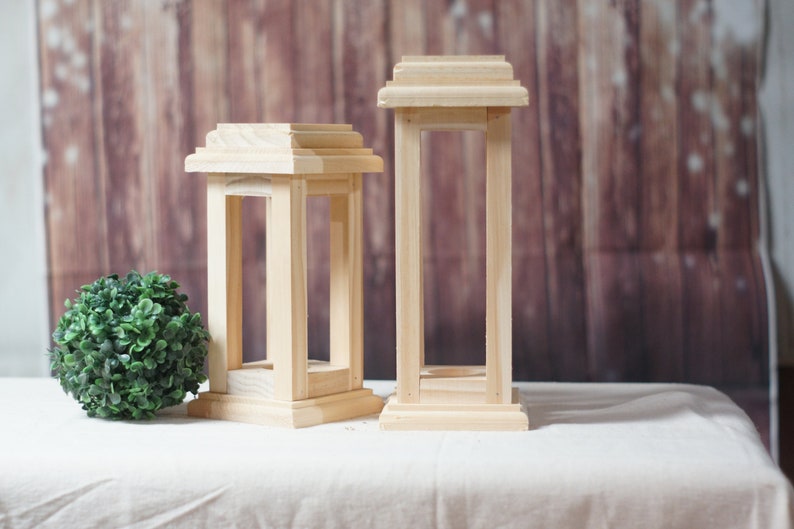 May include: Two wooden candle lanterns, one taller than the other, with a simple, rectangular design. The lanterns are made of unfinished wood and have a rustic look.