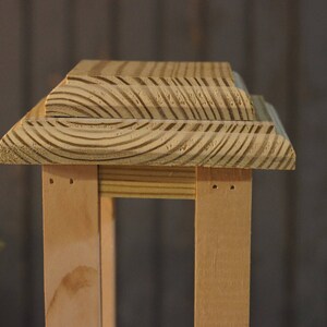 May include: A wooden lantern with a natural wood finish. The lantern has a square base and a rectangular top with a decorative, layered design.